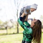Mother joyfully lifts her baby outdoors in a bright, sunny setting.
